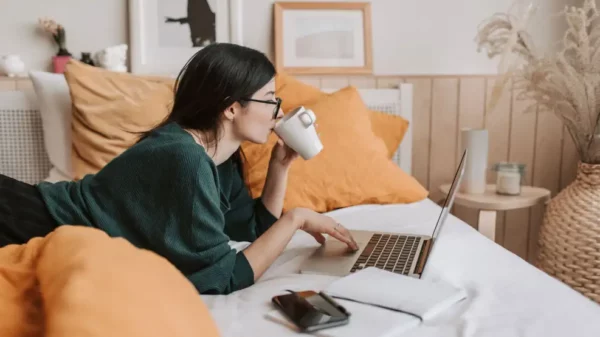 Mujer tomando café enfrente a un computador, recostada en la cama.